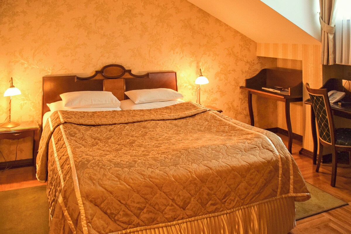 A bedroom with a large bed, a desk, and a chair. There are two bedside lamps and a window with curtains.
