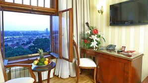 A hotel room with a large window offering a city view, a wooden desk with a telephone, a chair, and a small table with fruits. - Moskva (Belgrade)