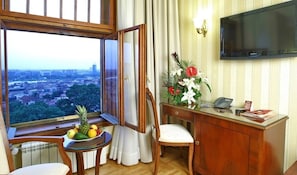 Habitación de hotel con un ventanal grande que ofrece vistas a la ciudad, un escritorio de madera con teléfono, una silla y una mesita con frutas.