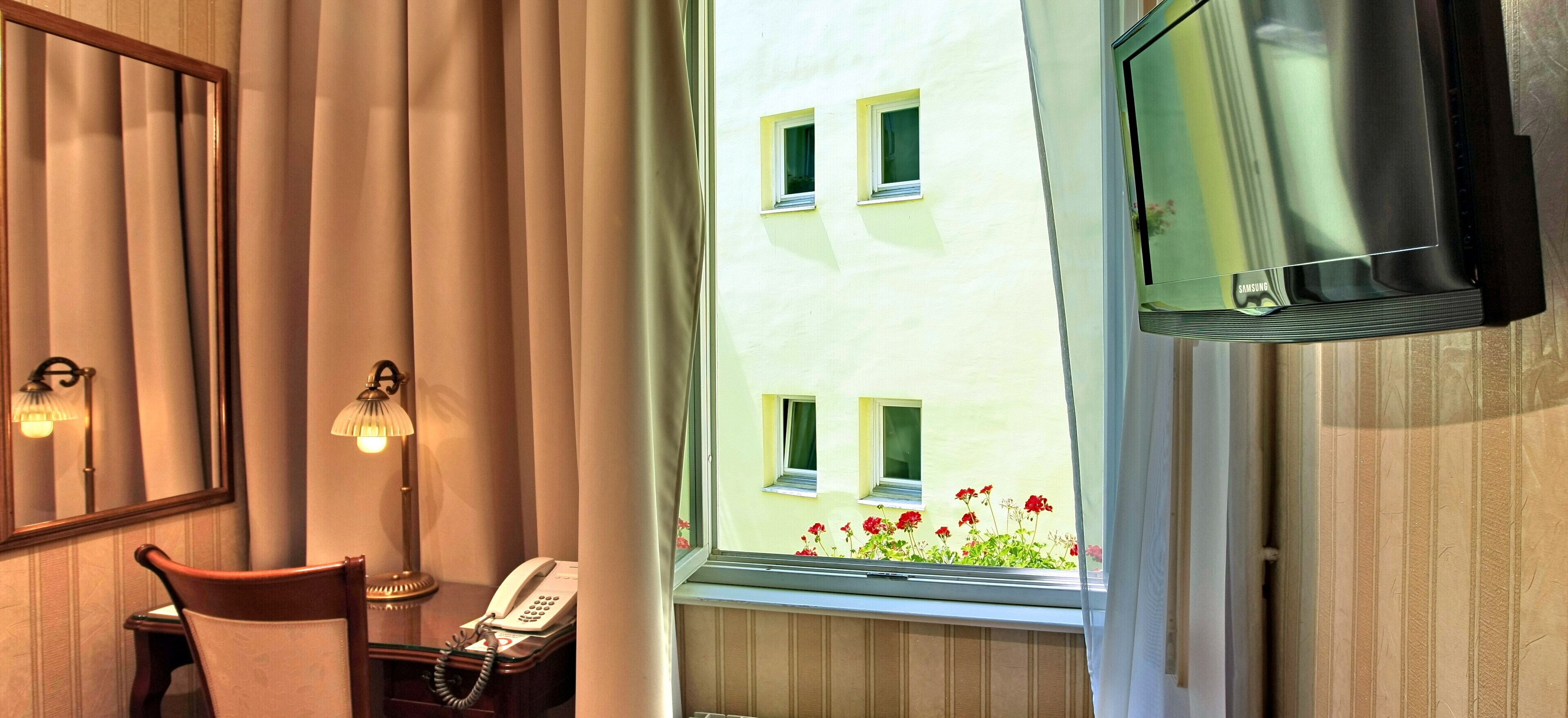 A hotel room with a bed, a bedside telephone, a window with curtains, a television mounted on the wall, and a view of a building with windows and flowers outside.