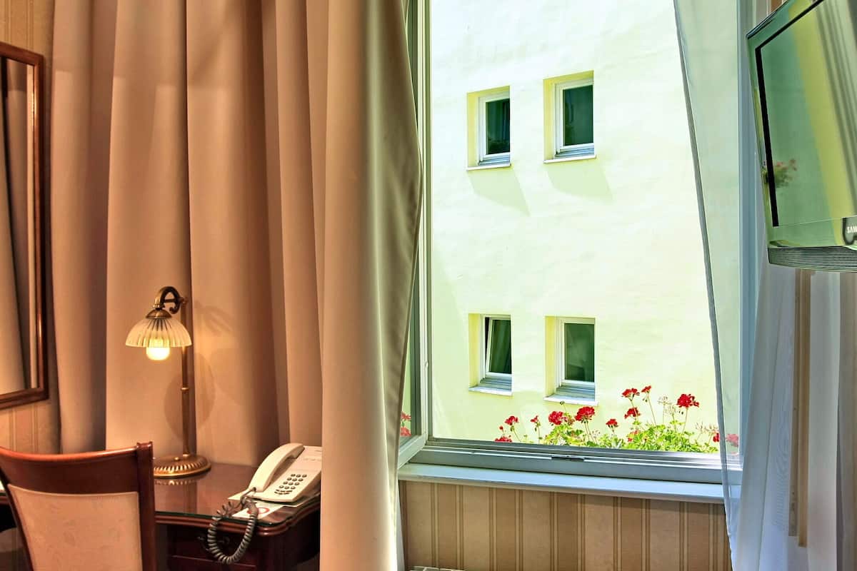 A hotel room with a bed, a bedside telephone, a window with curtains, a television mounted on the wall, and a view of a building with windows and flowers outside.