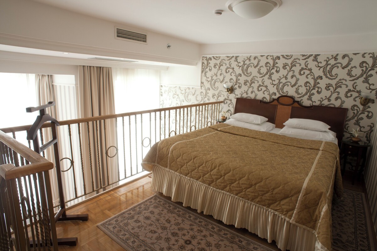 A bedroom with a large bed, a wooden railing, a patterned rug, and a window with curtains.