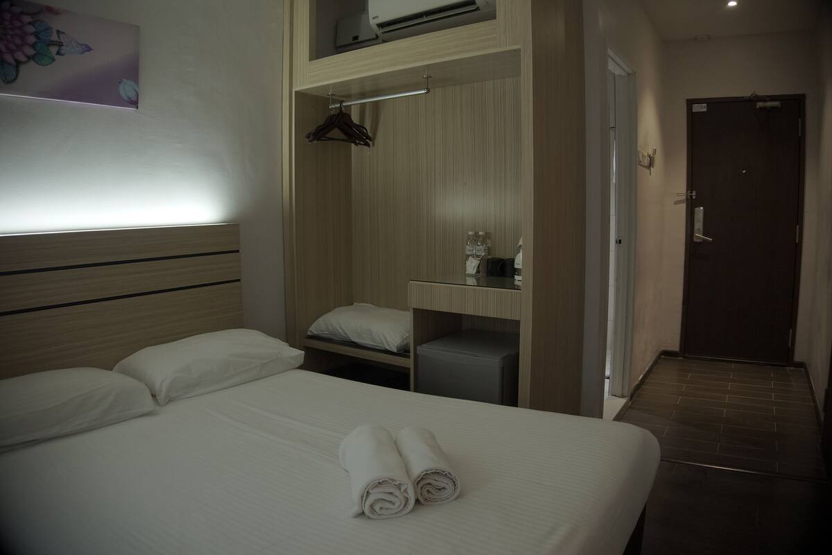 Quarto de hotel com cama, mesa de cabeceira, espelho, cabideiro e porta.
