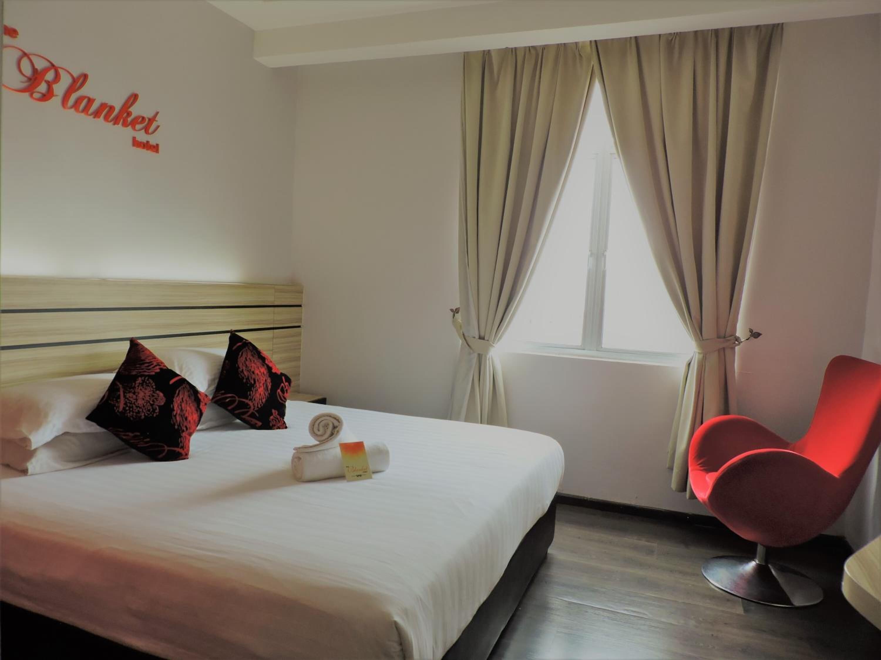 A hotel room with a bed, pillows, a red chair, a window with curtains, and a wall sign.