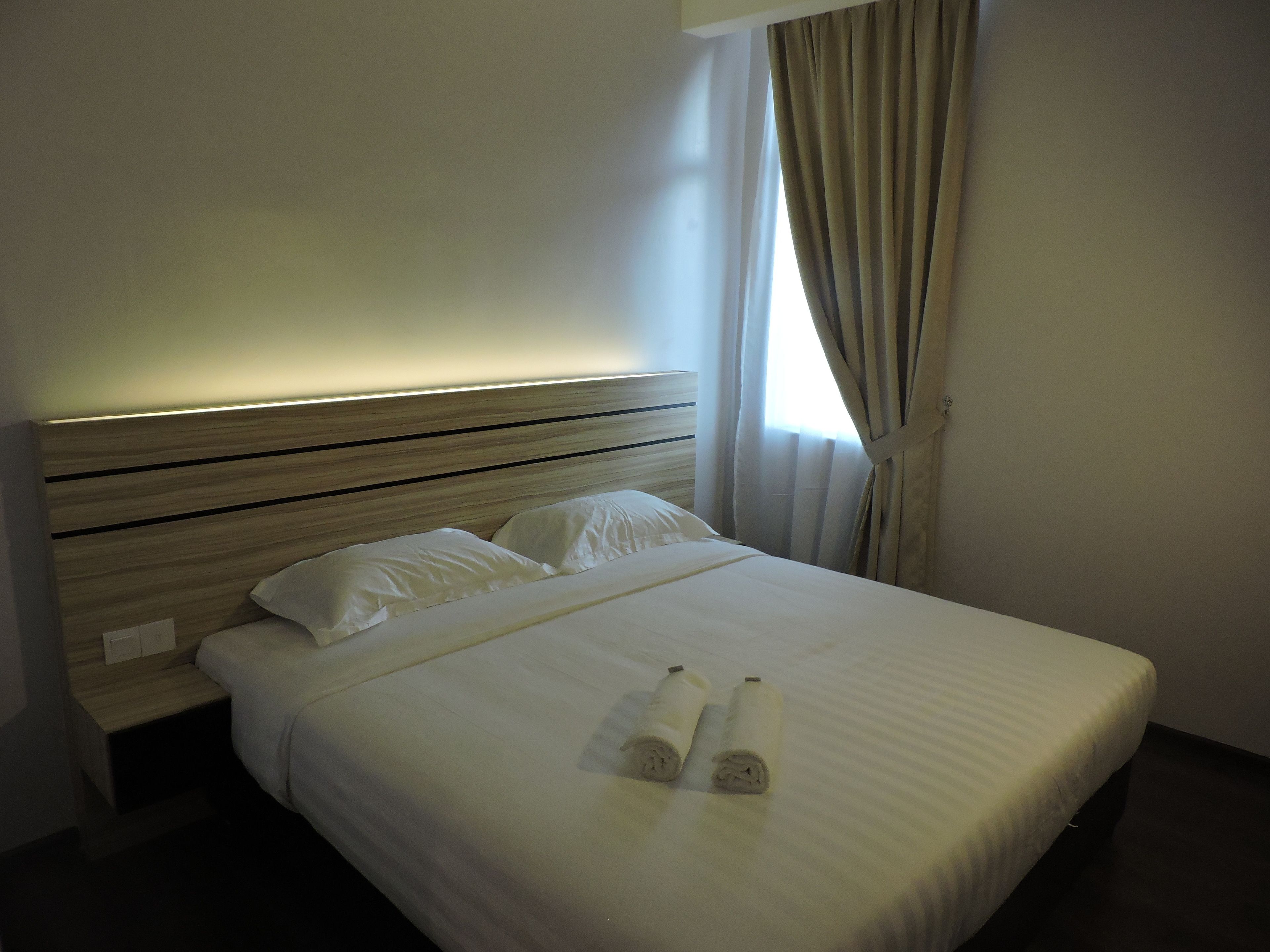 A neatly made bed with white linens and two rolled towels, a wooden headboard, and a window with curtains.