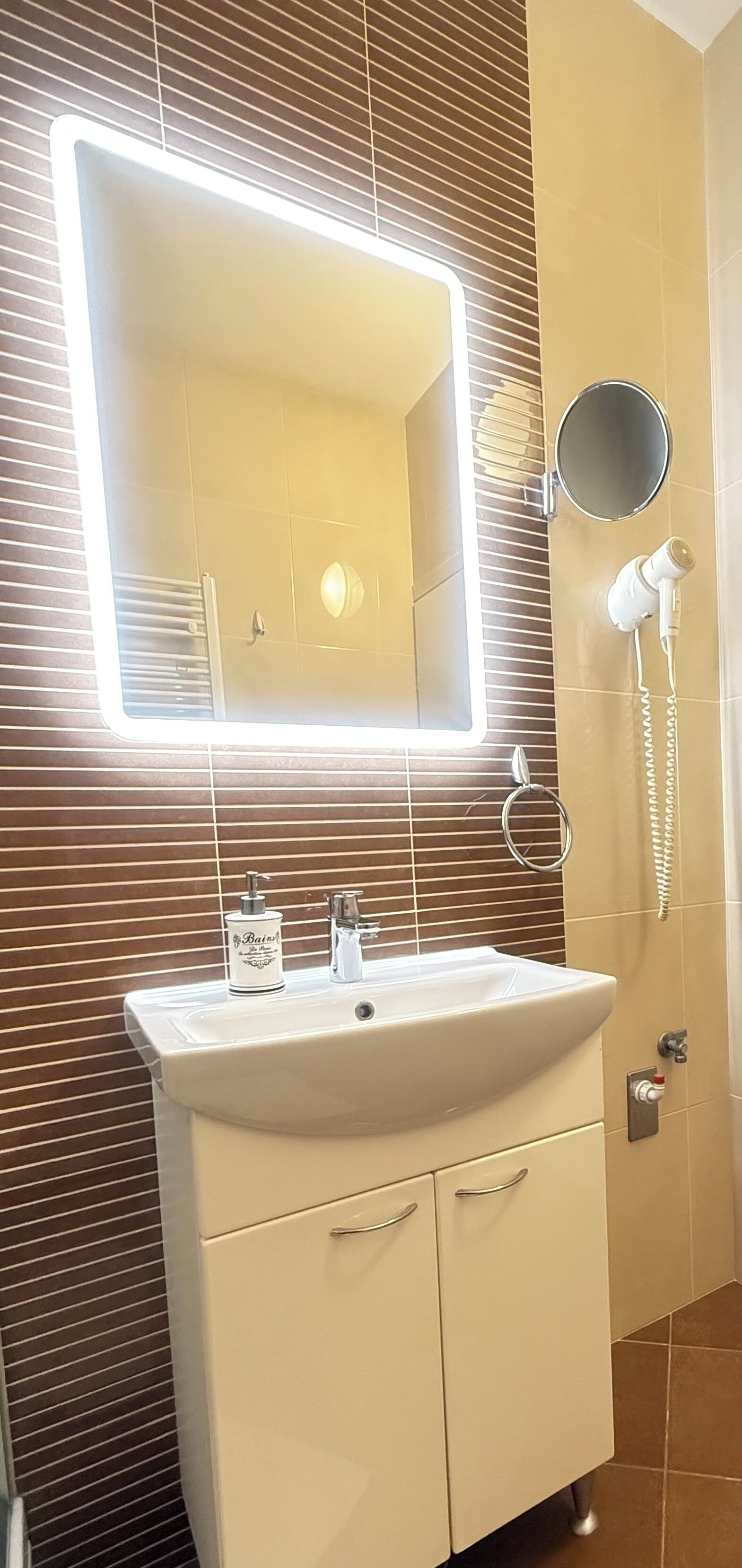 Un baño con lavabo blanco, un espejo con luz, un secador de pelo y un aro para toallas.