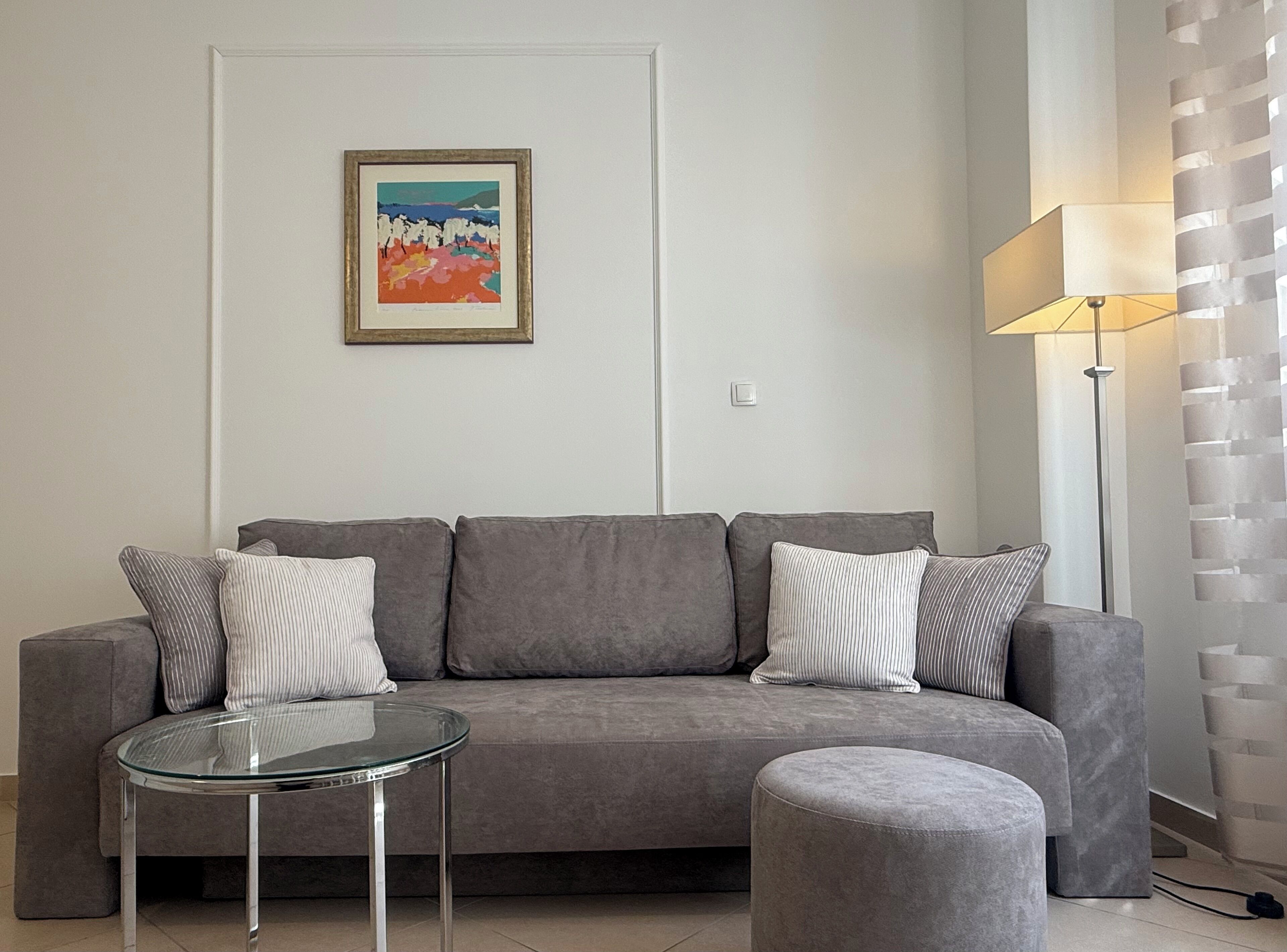 A modern living room with a grey sofa, a glass coffee table, and a grey ottoman. A framed painting is on the wall, and a floor lamp is to the right.