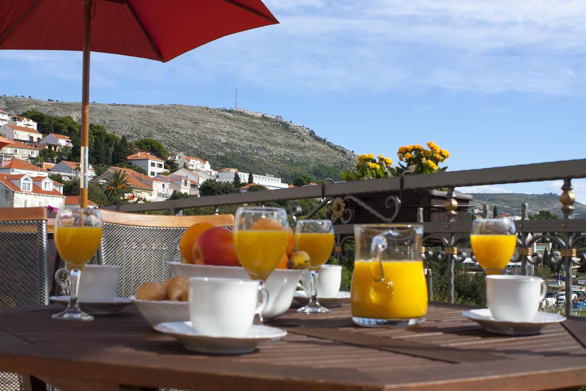 Mesa de café da manhã com suco de laranja, xícaras de café e doces, com vista para uma colina com casas.