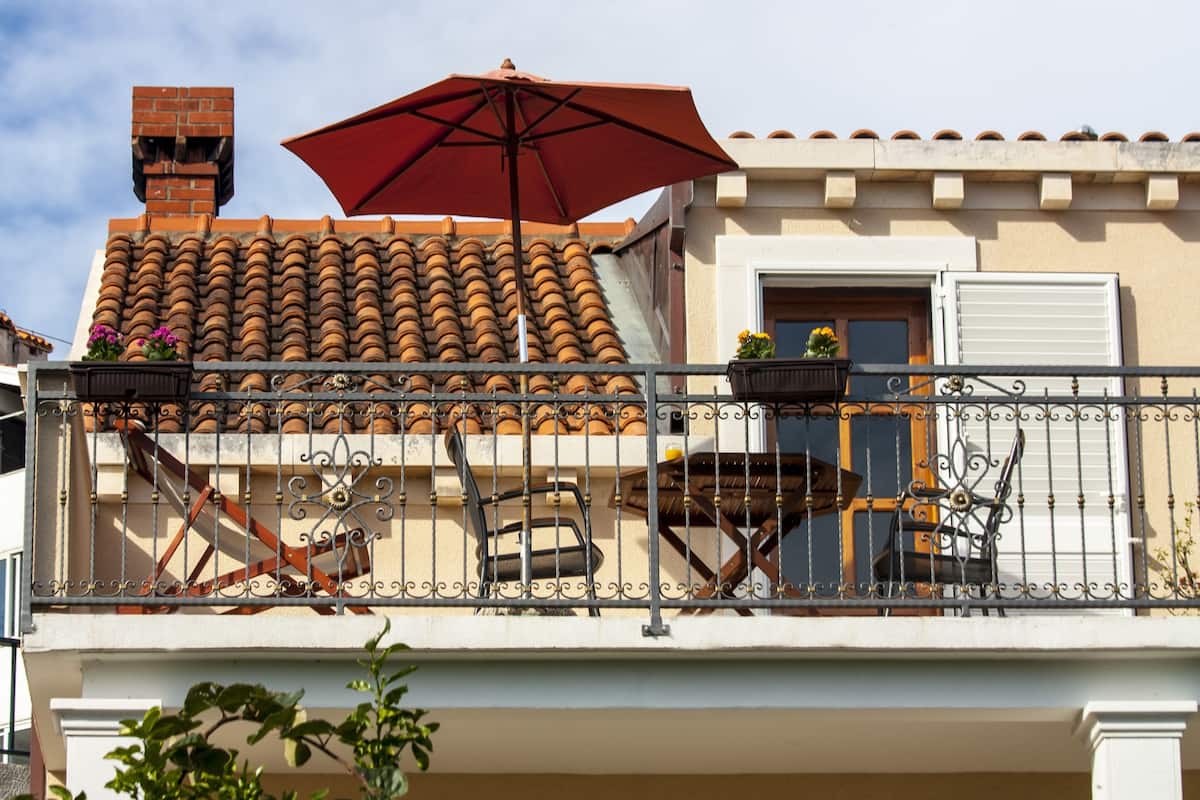 Uma varanda com um guarda-sol vermelho, telhado de azulejos e mobiliário de exterior.