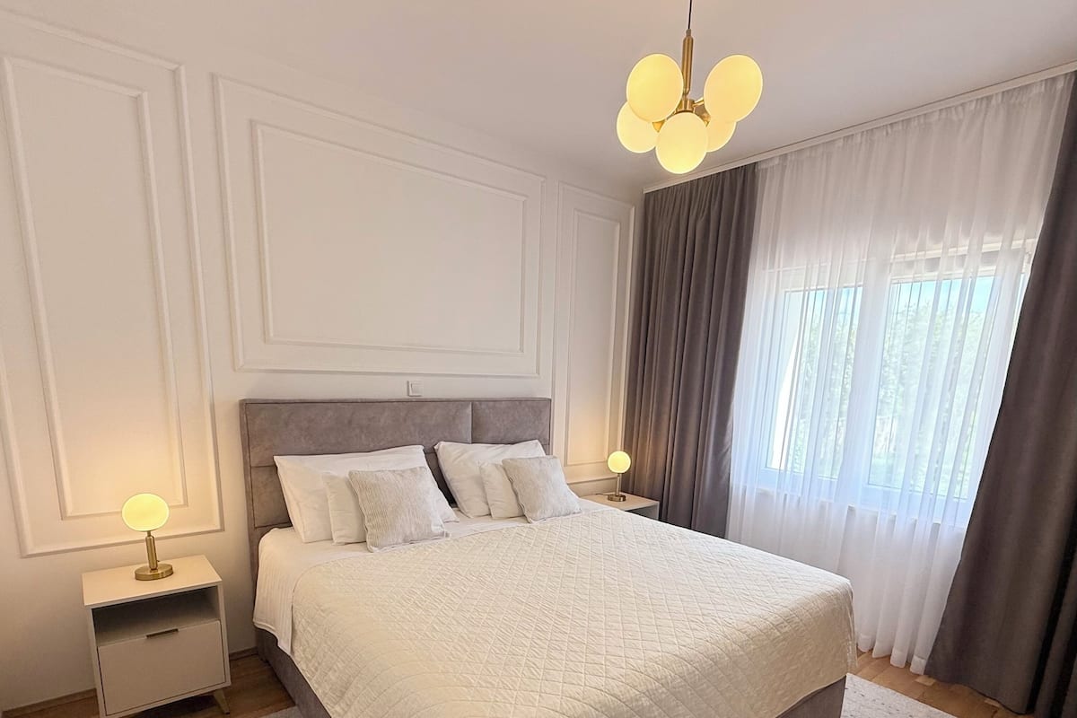 Quarto com uma cama grande, luminárias de cabeceira, mesa de cabeceira e uma janela com cortinas.