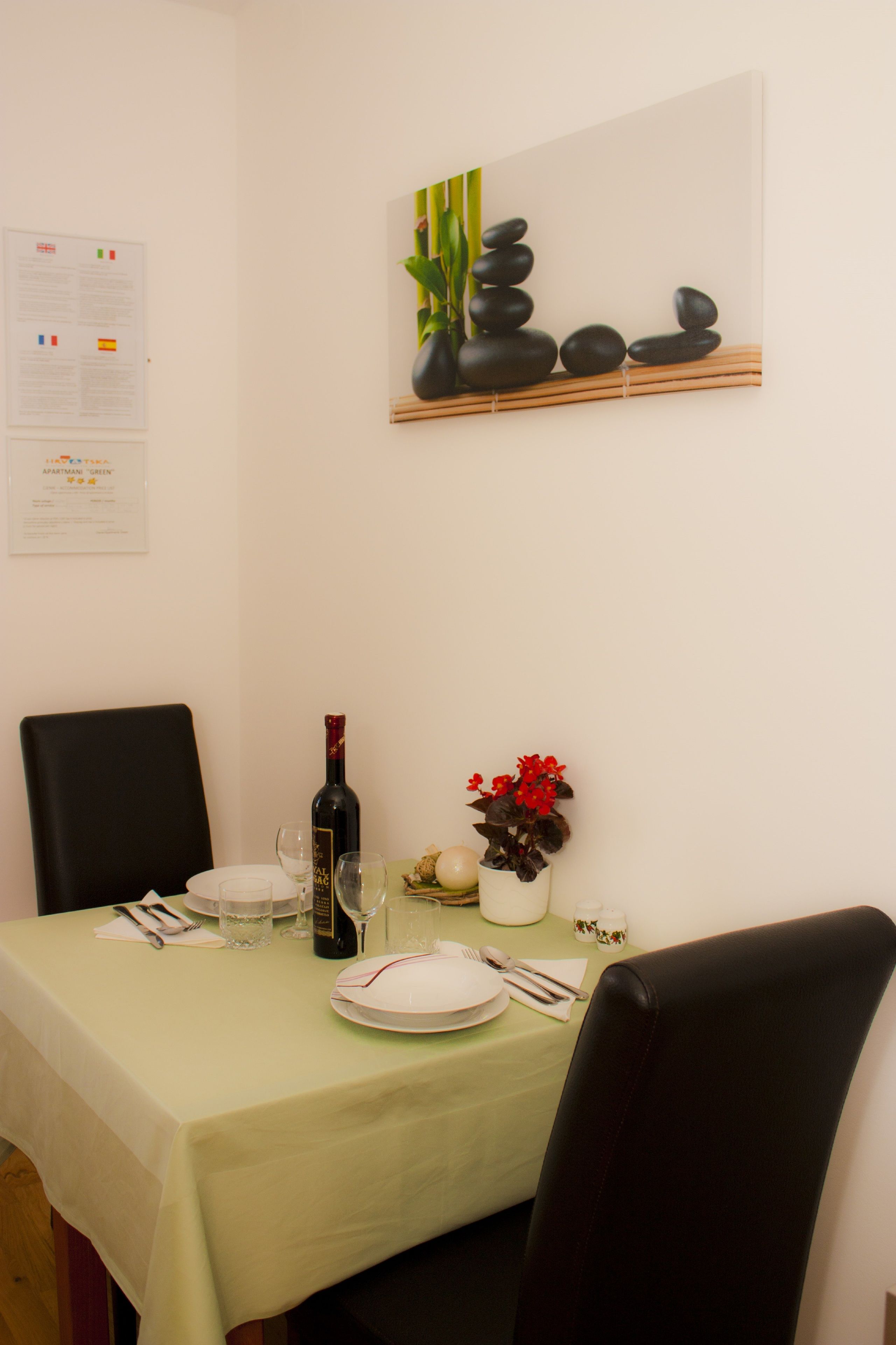 A dining table set with plates, cutlery, and a bottle of wine, with a vase of red flowers and a painting of stones and greenery on the wall.