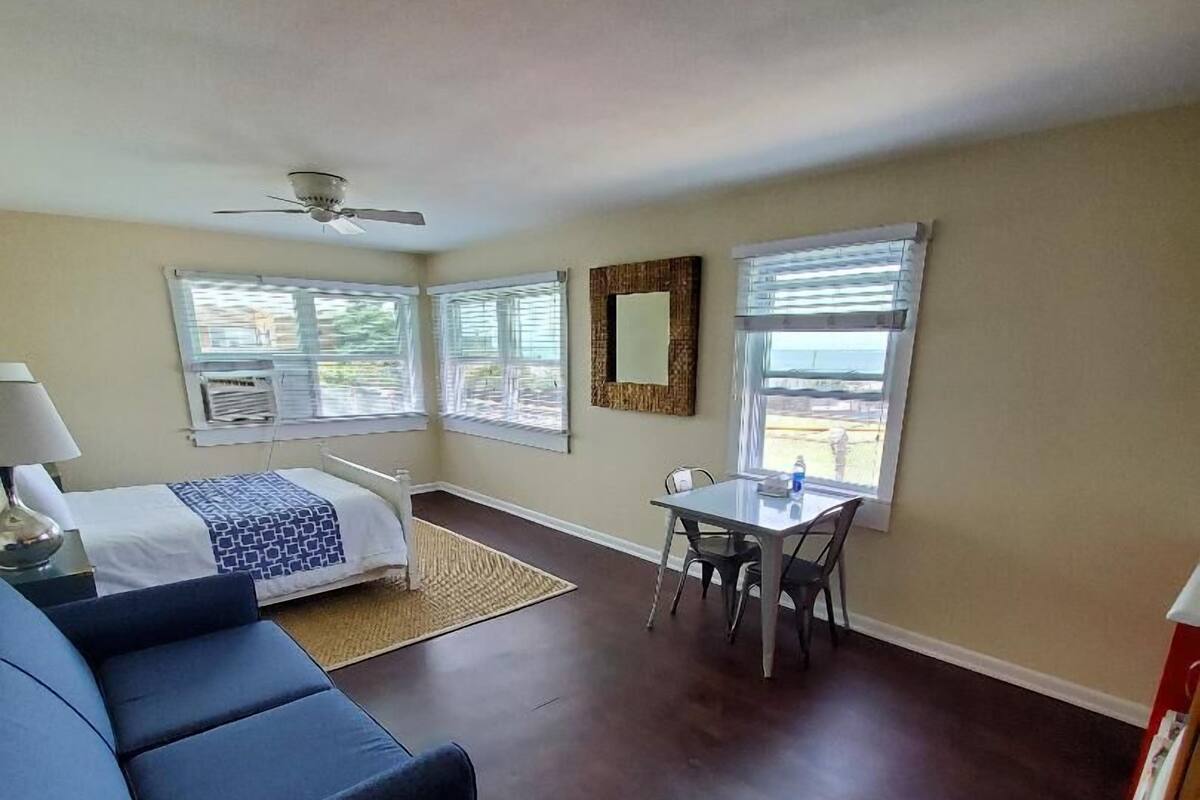 A room with a bed, a blue sofa, a dining table with chairs, and a ceiling fan.
