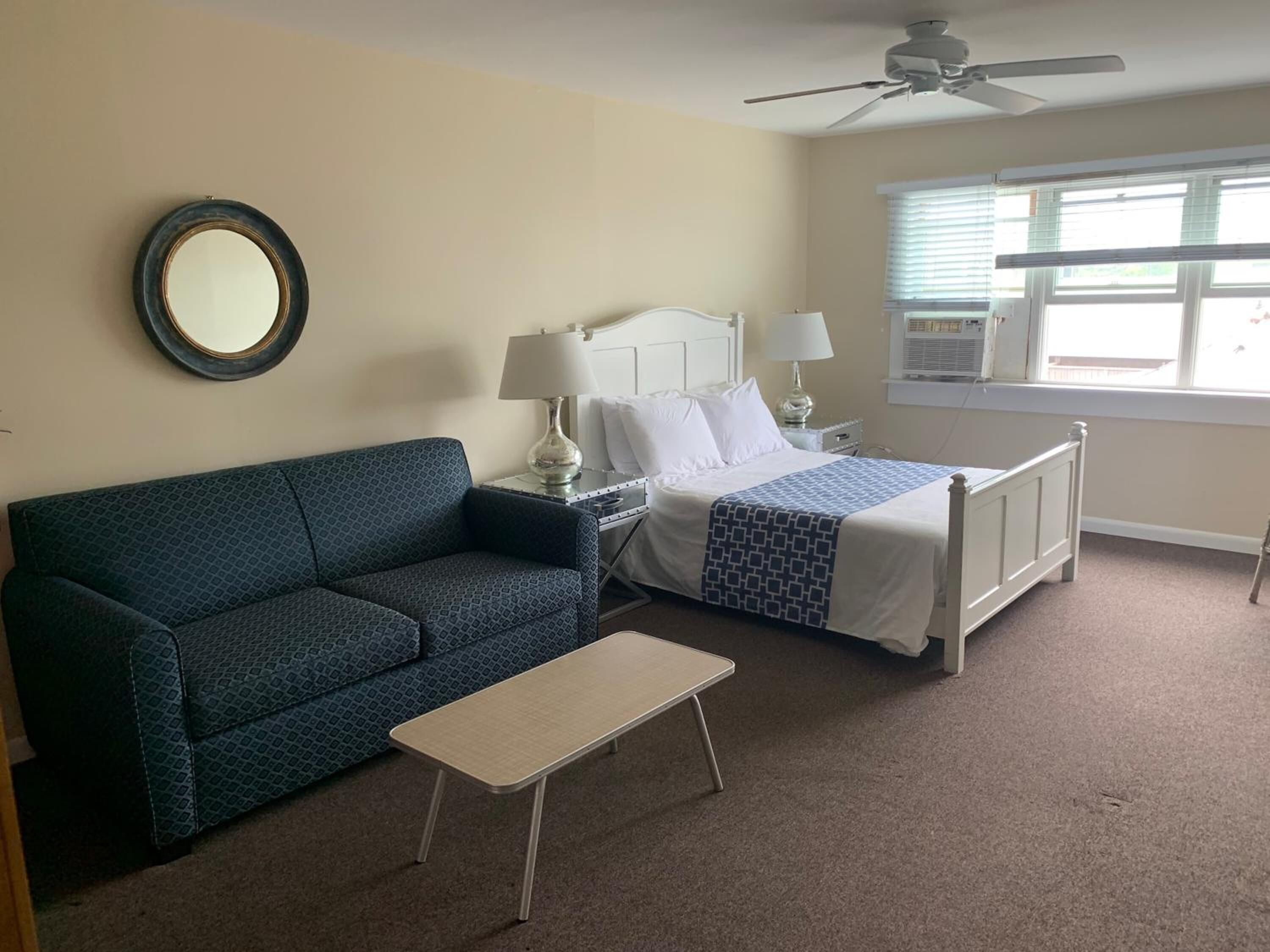 A hotel room with a bed, a sofa, a small table, a ceiling fan, and a window with blinds.