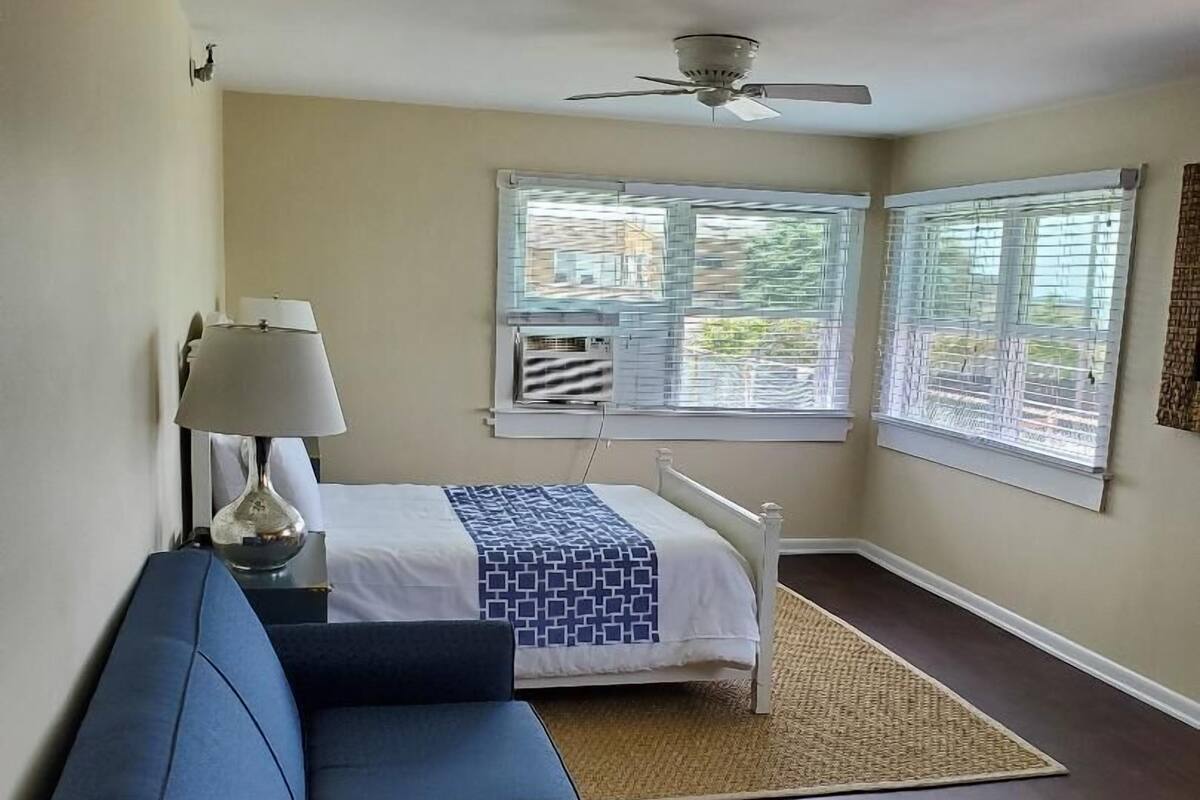 A bedroom with a bed, a ceiling fan, a window with blinds, a nightstand with a lamp, and a dark-colored sofa.
