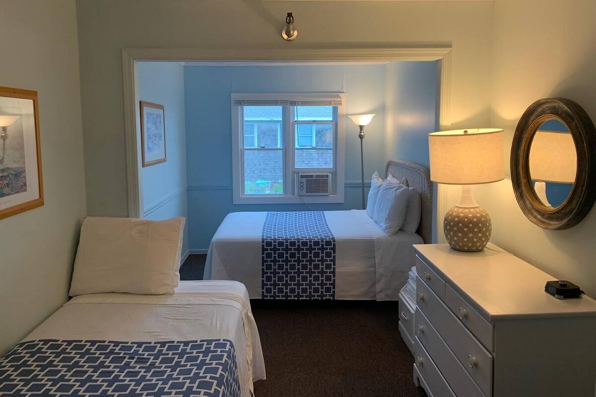 A bedroom with two beds, a dresser, a lamp, and a window with an air conditioning unit.