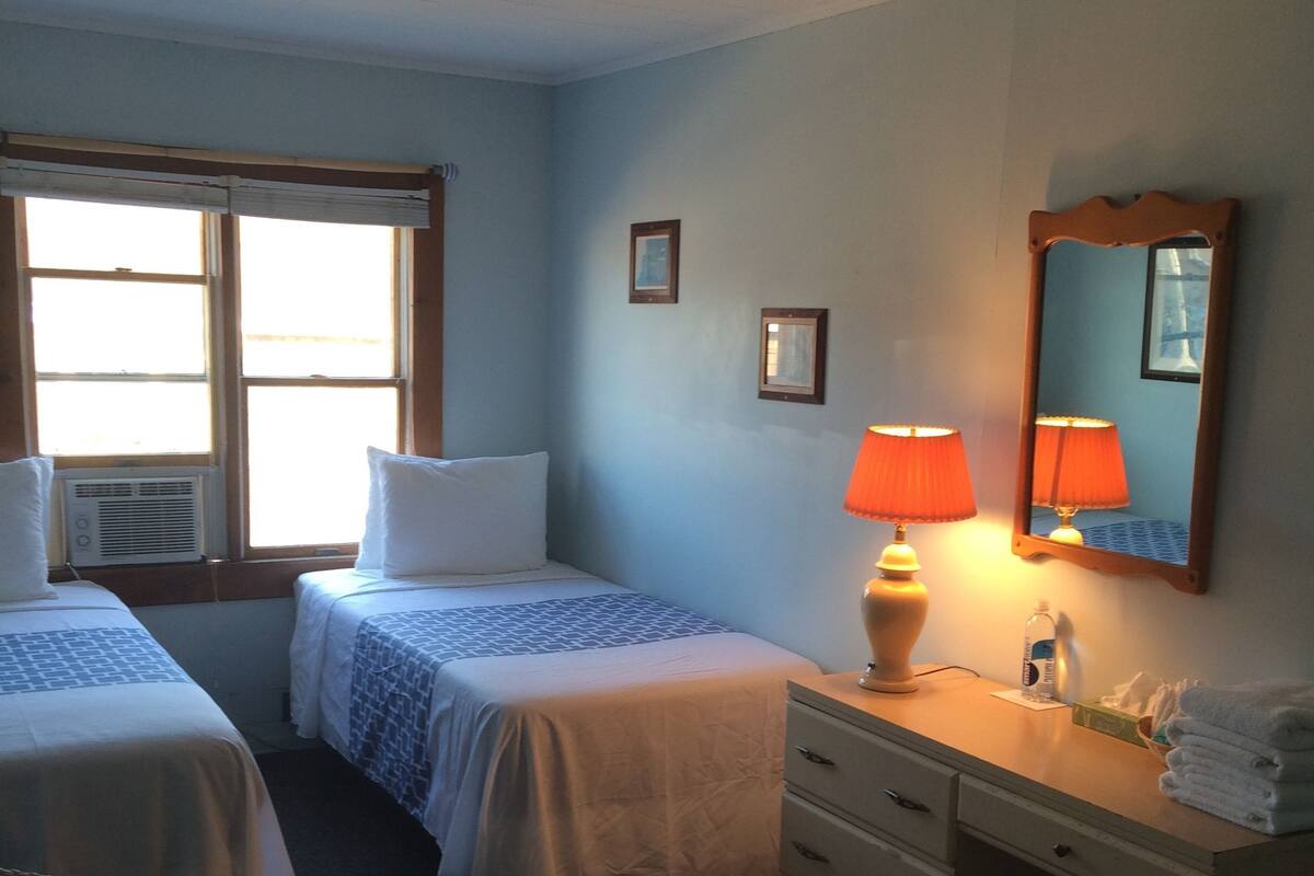 A hotel room with two beds, a desk with a lamp, a mirror, and a window with blinds.