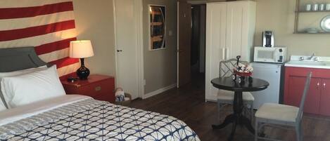 Une chambre d’hôtel avec un grand lit, un bureau avec une chaise, une petite kitchenette et une porte donnant sur une autre pièce.