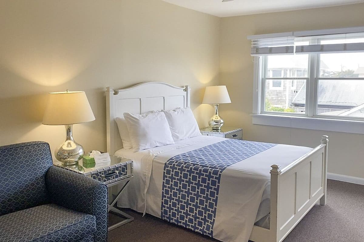 A hotel room with a bed, two bedside lamps, a chair, a window with blinds, and a view of buildings outside.