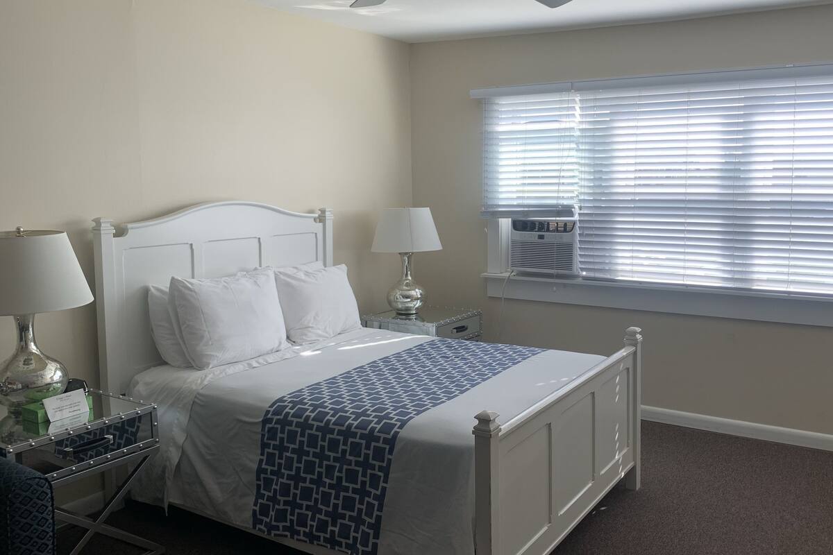 A bedroom with a bed, two bedside tables, a lamp, a window with blinds, and a ceiling fan.