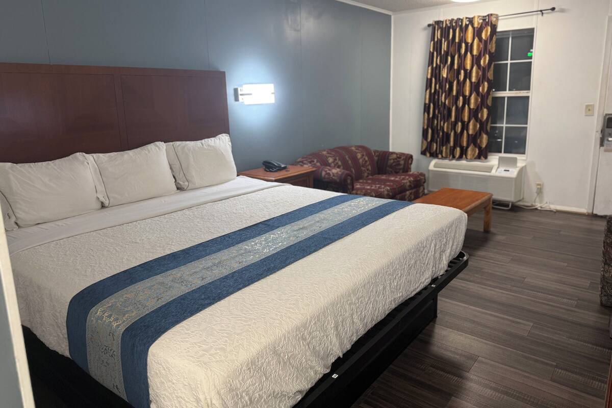 Una habitación de hotel con una cama grande, cabecera de madera, una mesita pequeña, un sillón con estampado, una ventana con cortinas y un equipo de aire acondicionado de pared.