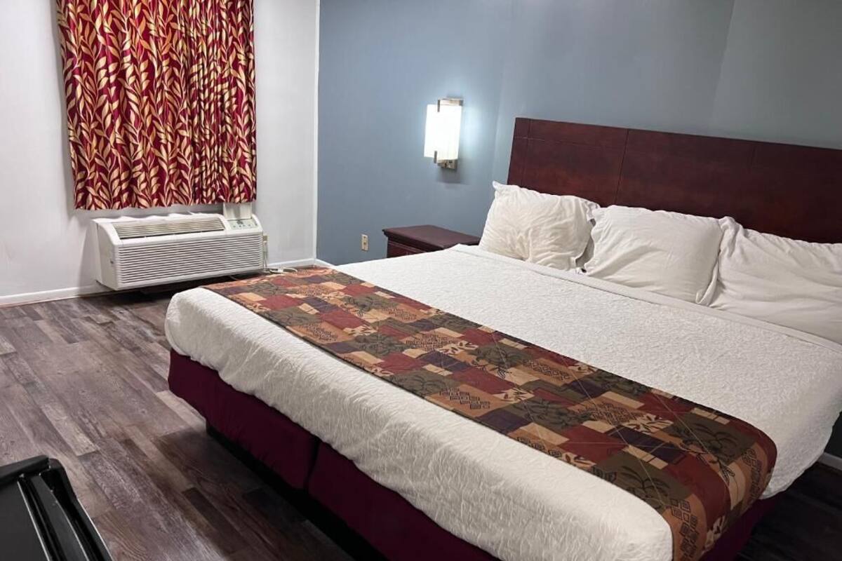 Una habitación de hotel con una cama grande, una colcha roja y blanca estampada, un calentador de pared y una mesita de noche con una lámpara.