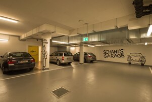 Um estacionamento com vários carros, uma porta amarela com a inscrição "Hotel" e uma parede com a inscrição "SCHANN'S GARAGE" e um desenho de carro.