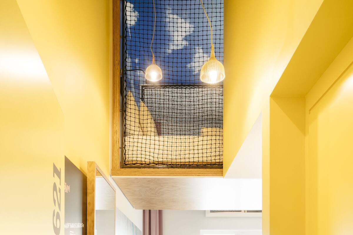 A modern hotel room with a yellow color scheme, a wooden ceiling, a bed with a view, a desk with a chair, and a window with curtains.