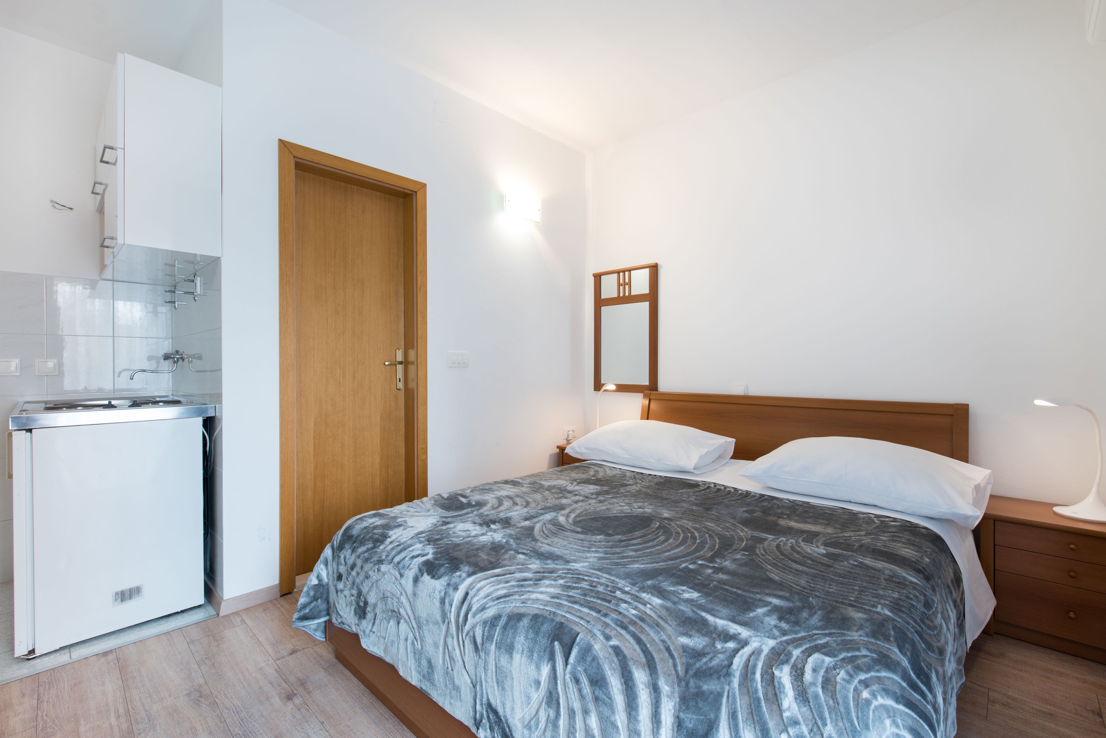 A bedroom with a bed, bedside table, and a small kitchenette area.