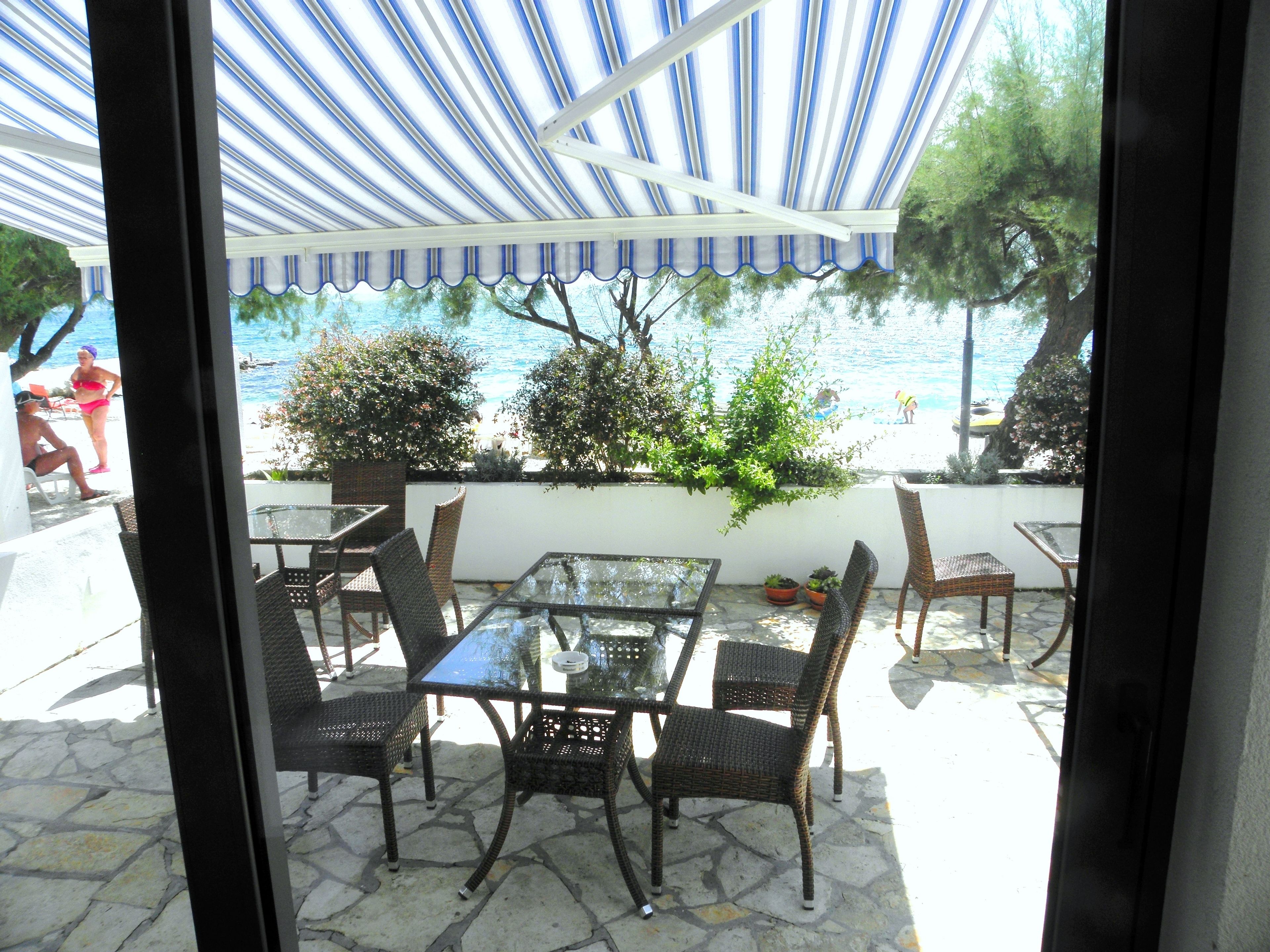 An outdoor patio with a glass table and wicker chairs, overlooking a beach and the sea.