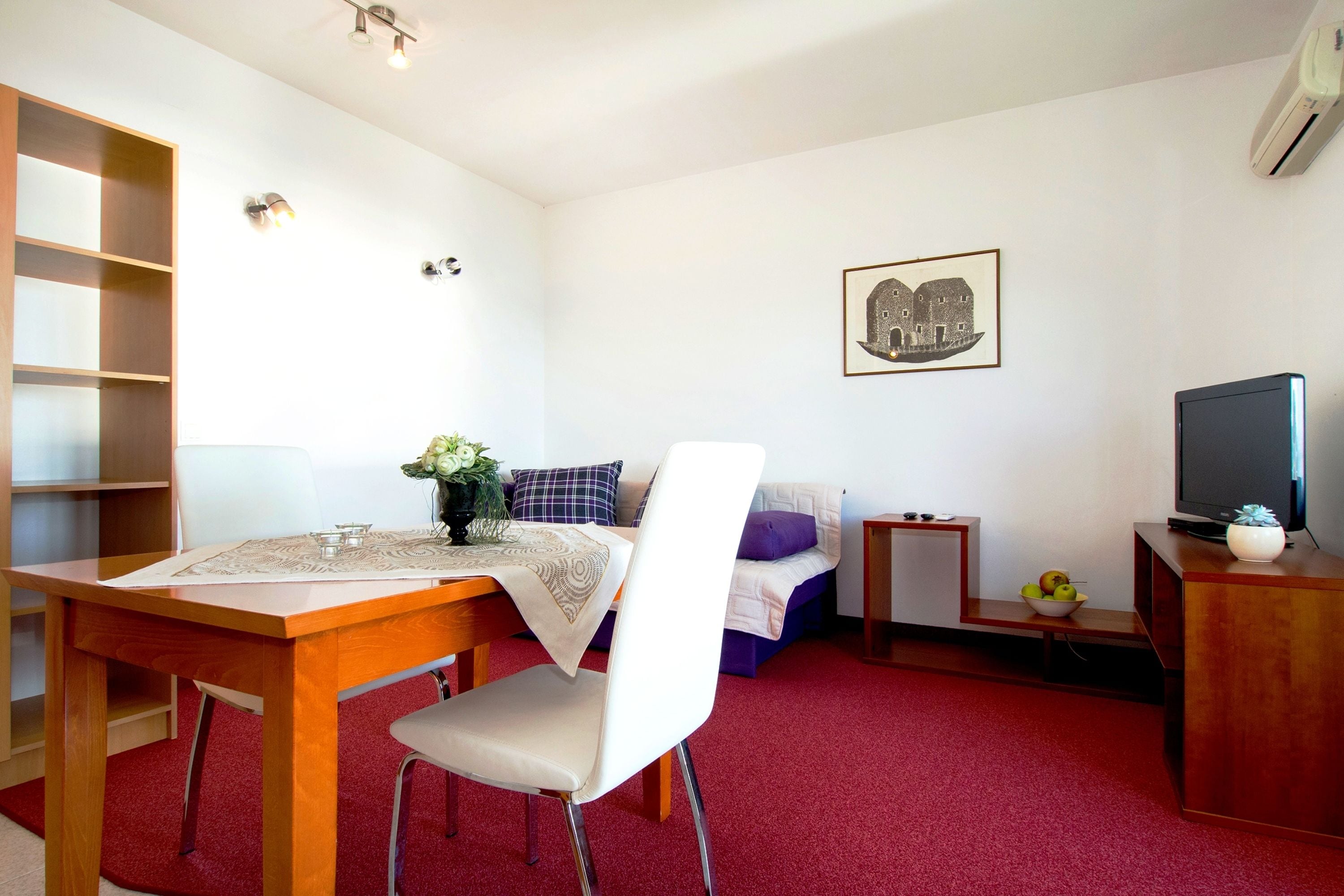 Um espaço compacto com uma mesa de jantar, uma cadeira, uma cama com travesseiro roxo, uma mesinha de cabeceira com uma tigela de frutas e uma televisão em um suporte de madeira.