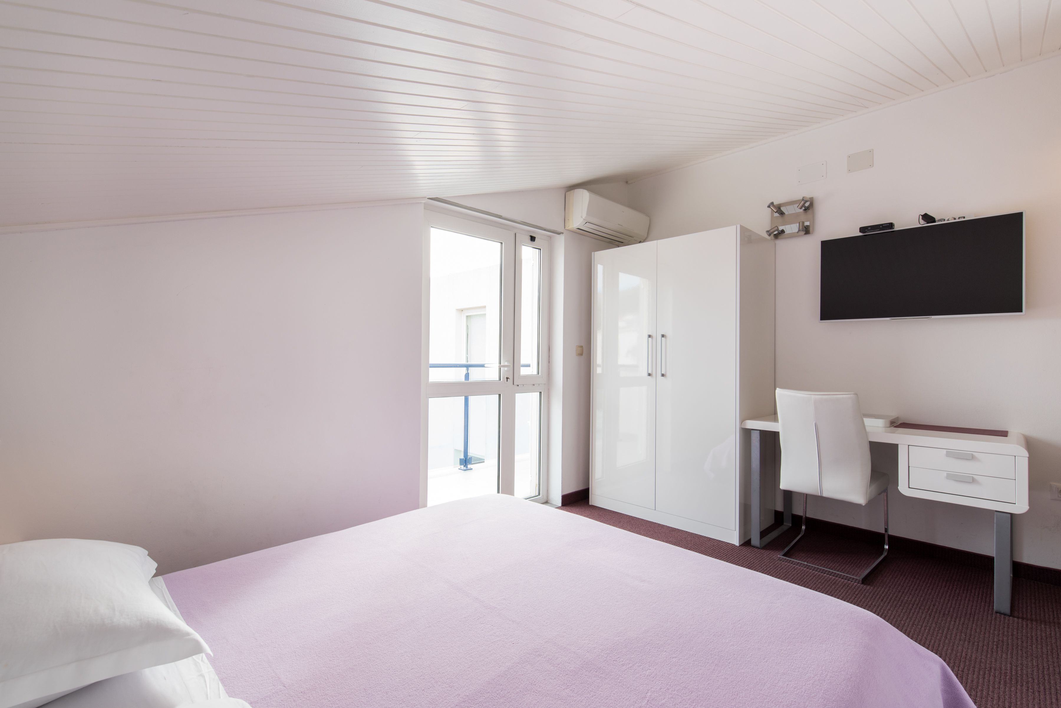 A bedroom with a bed, a desk, a chair, a wardrobe, and a TV mounted on the wall.