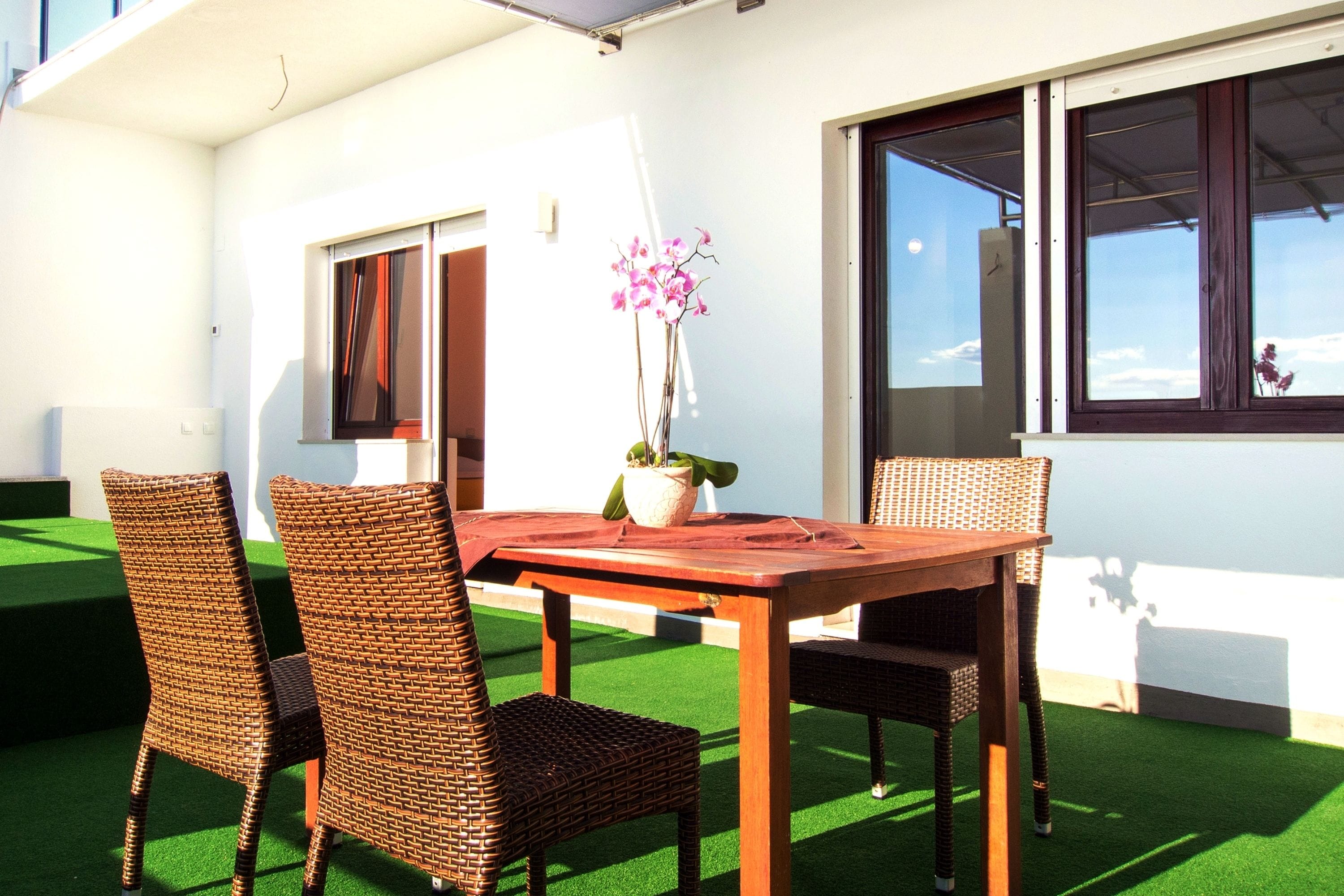 A patio with a wooden table and wicker chairs, a potted plant, and a view of the sky through a window.