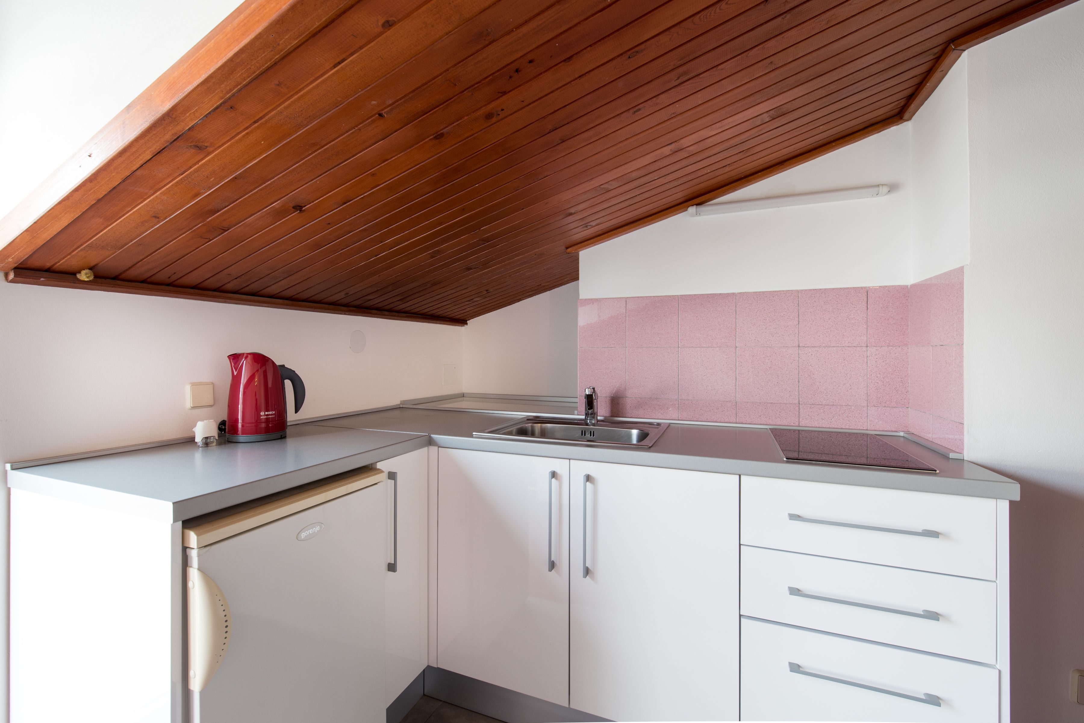 A compact kitchen with white cabinets, a stainless steel sink, and a red kettle.