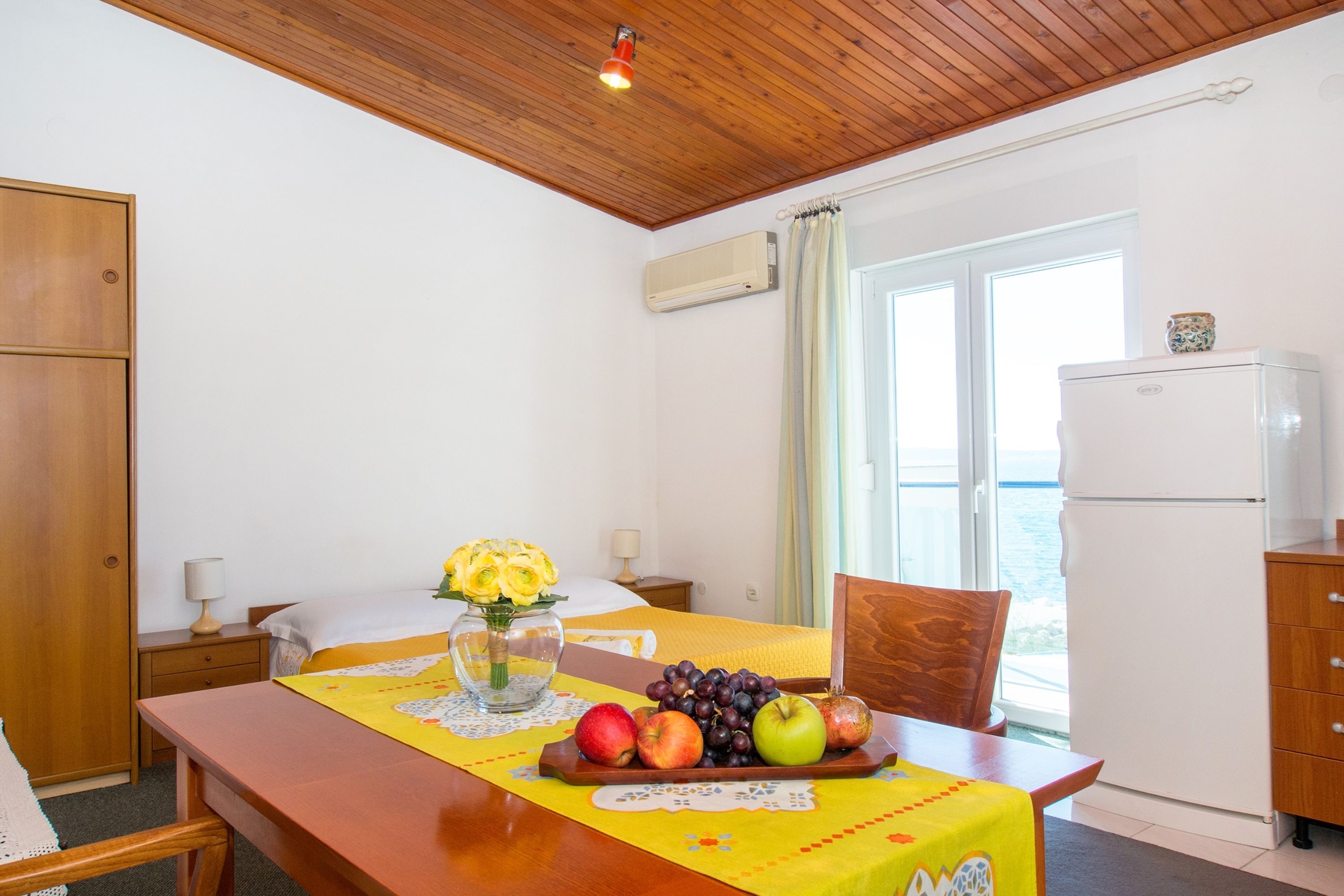 A hotel room with a wooden ceiling, a bed with a yellow bedspread, a dining table with fruits and a vase of flowers, a refrigerator, and a window with a view of the sea.