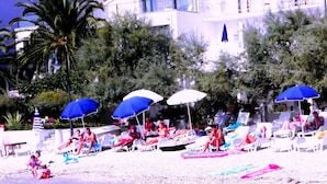 A beach with sun loungers and umbrellas, a palm tree, and a building in the background. - Beachfront Villa Mediteran (Podstrana)