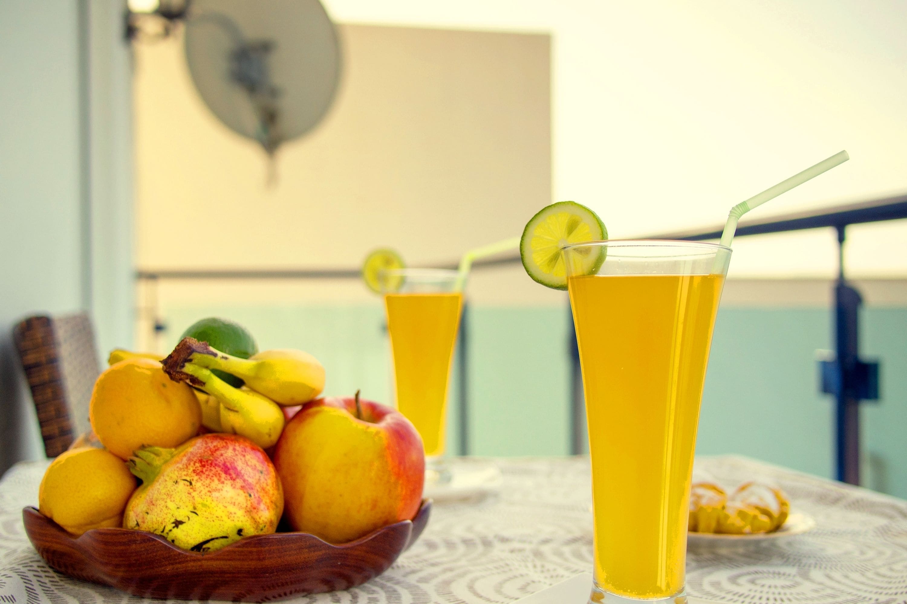 Una mesa con un frutero que contiene manzanas y cítricos, y dos vasos de jugo de naranja con pajitas y rodajas de lima.