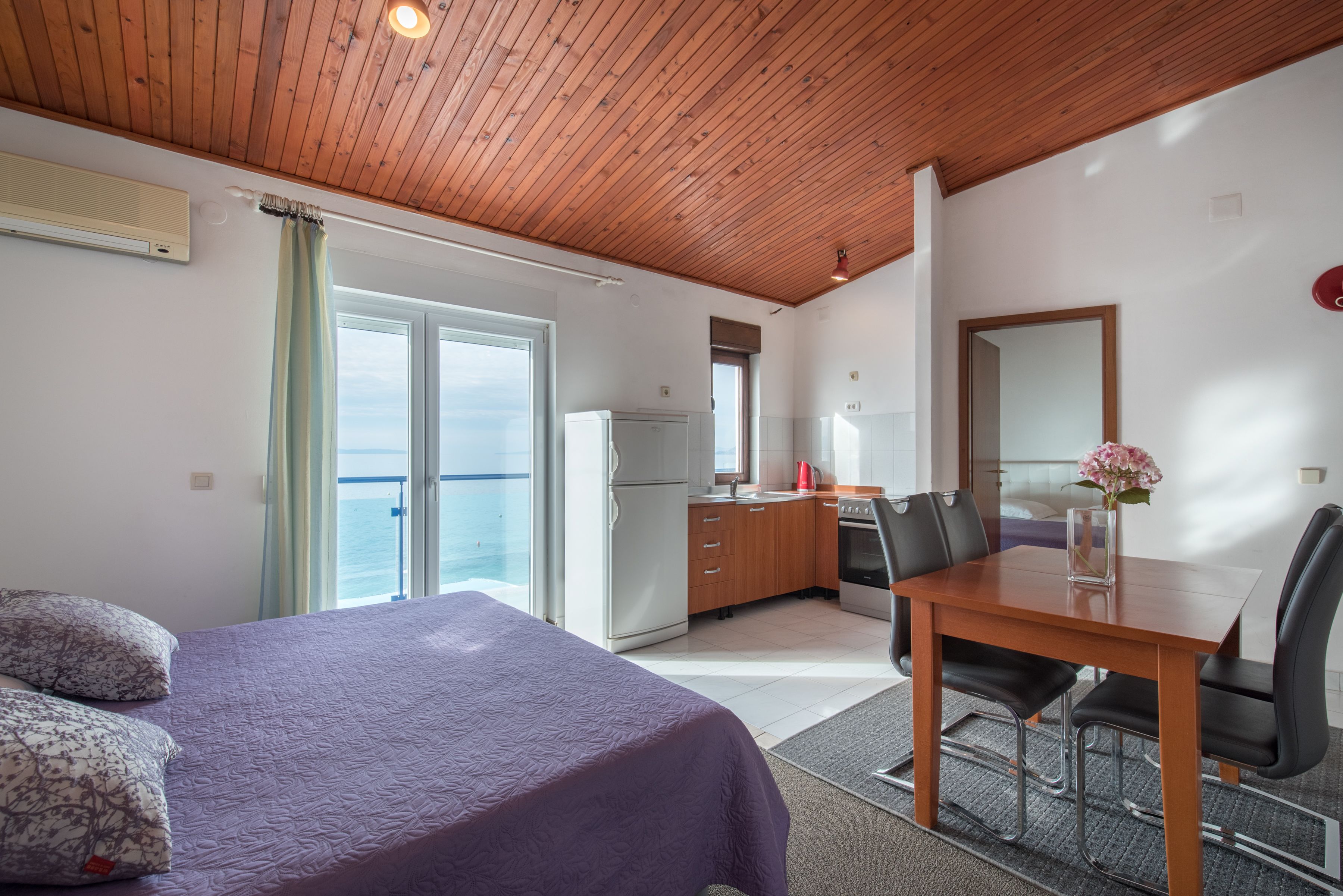 A bedroom with a bed, a desk, and a dining table with chairs. There is a view of the sea through the window.