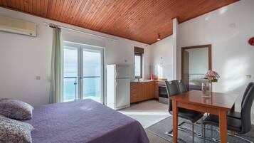 Quarto com cama, escrivaninha e mesa de jantar com cadeiras. Vista para o mar pela janela.