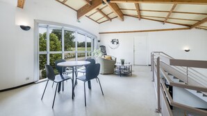 Una habitación amplia con techo alto y vigas de madera, que cuenta con una mesa redonda con sillas, un sofá y una pequeña estantería.