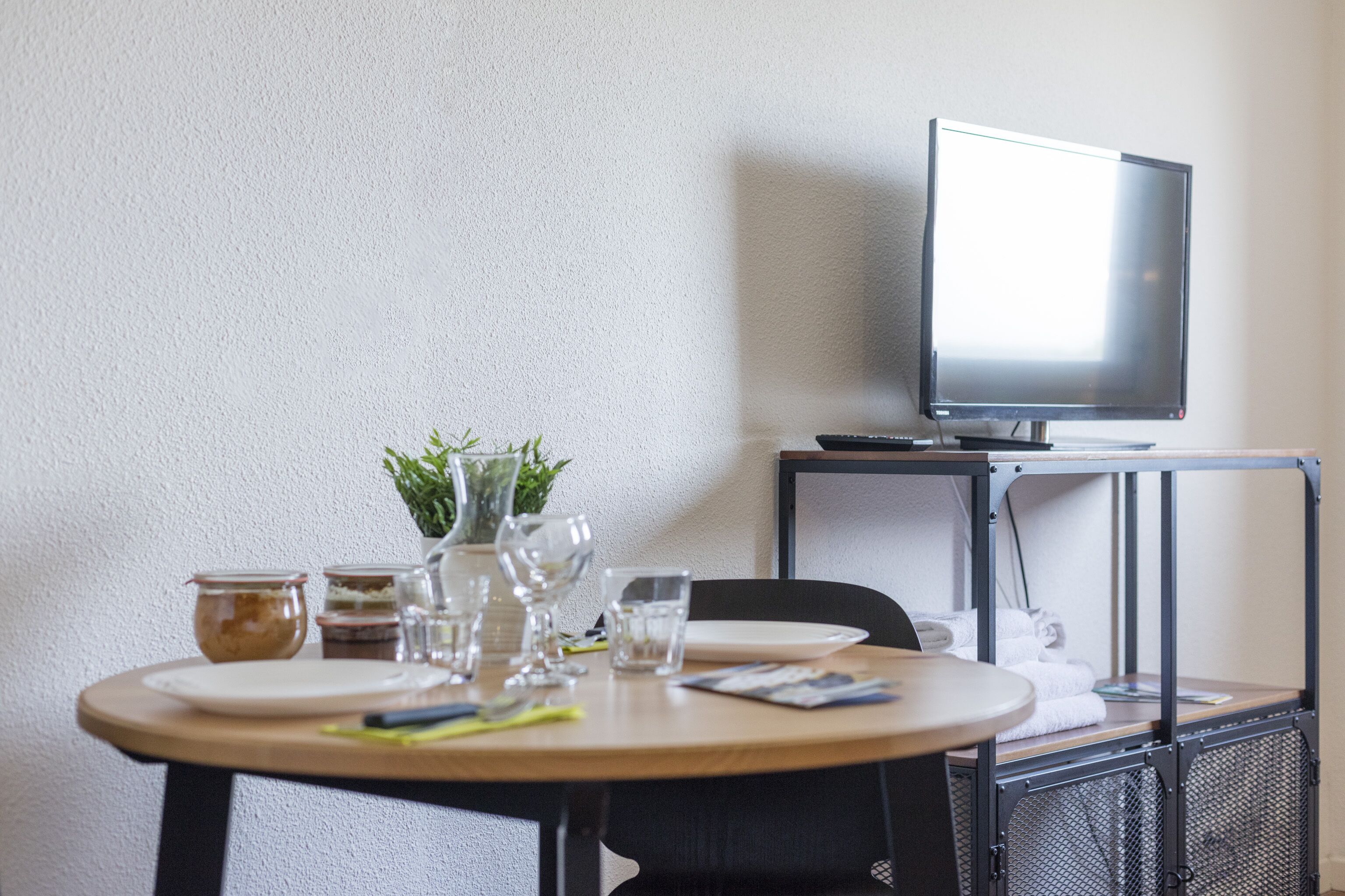 A room with a round table set for dining, a television on a stand, and a small plant on the table.