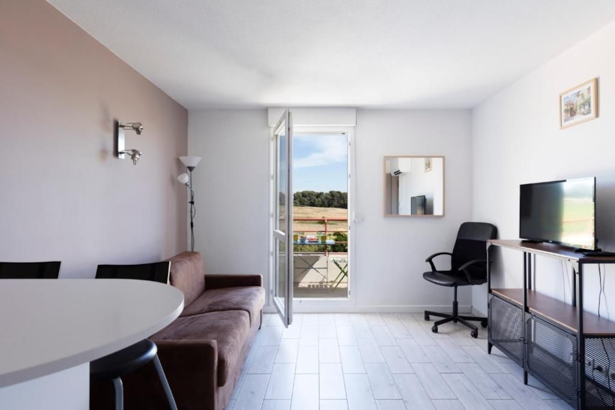 A modern room with a white table, brown sofa, a TV on a stand, a chair, and a door leading to an outdoor area.