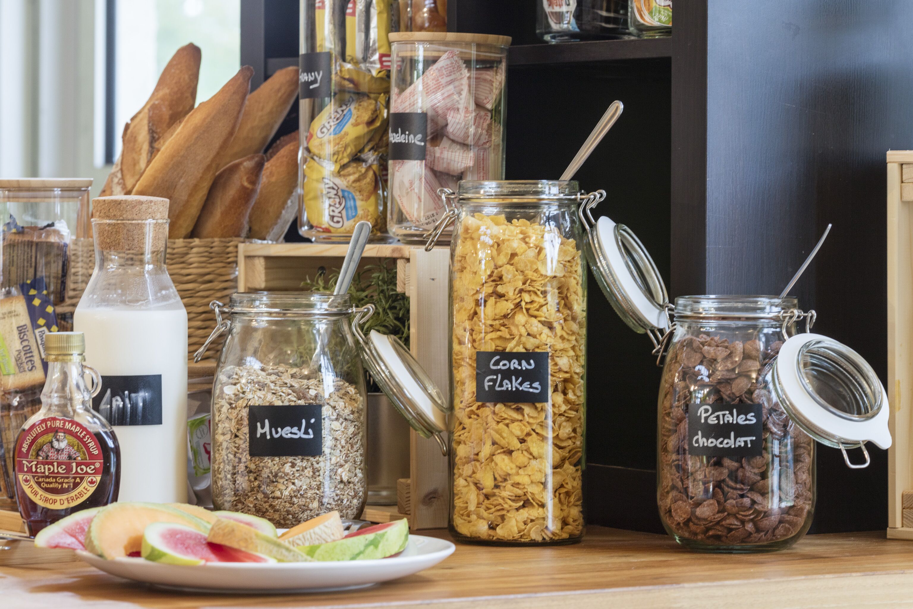 A variety of food items including jars labeled with contents like 'Huesli', 'Corn Flakes', and 'Petites chocolat', along with bread and a plate of sliced fruits.