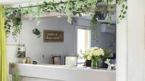 Un área de recepción con mostrador blanco, un cartel enmarcado, un jarrón con flores blancas y decoración colgante de hojas verdes.