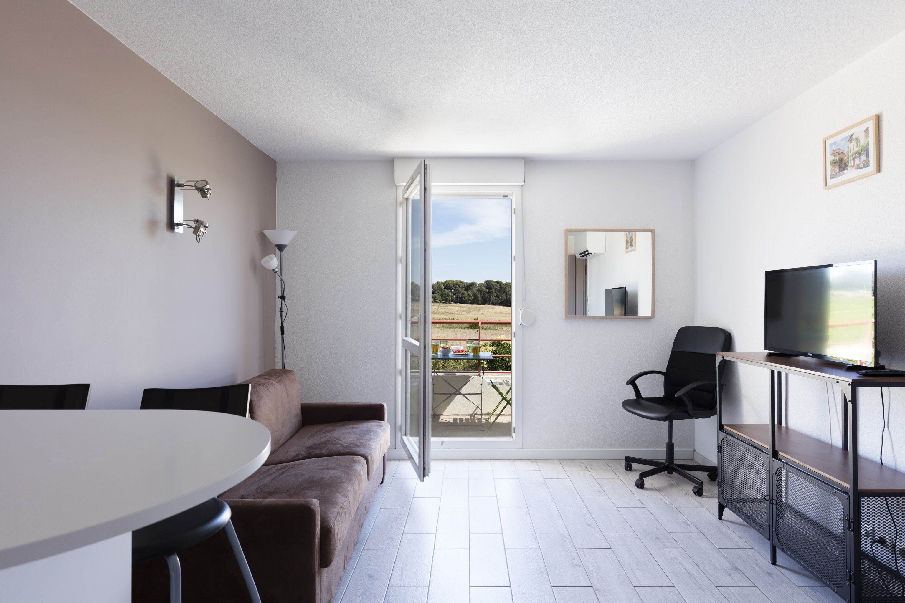 Una habitación moderna con una mesa redonda blanca, un sofá marrón, un televisor sobre un mueble de madera y un ventanal corredizo que da a un espacio exterior.