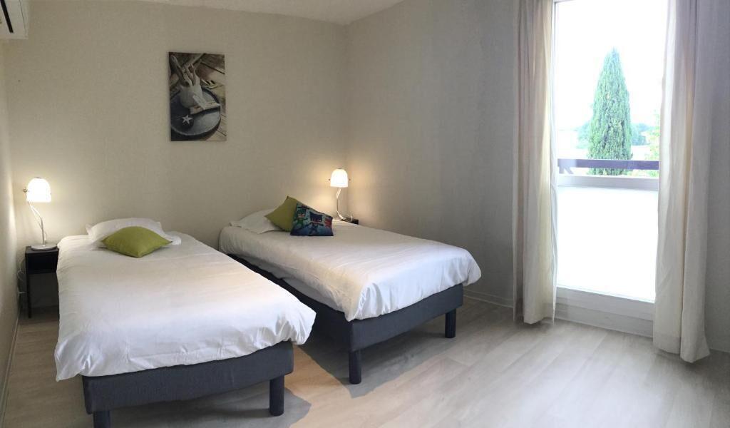 Una habitación con dos camas, cada una con lámpara y almohadas al lado, un cuadro en la pared y vistas a través de una ventana.