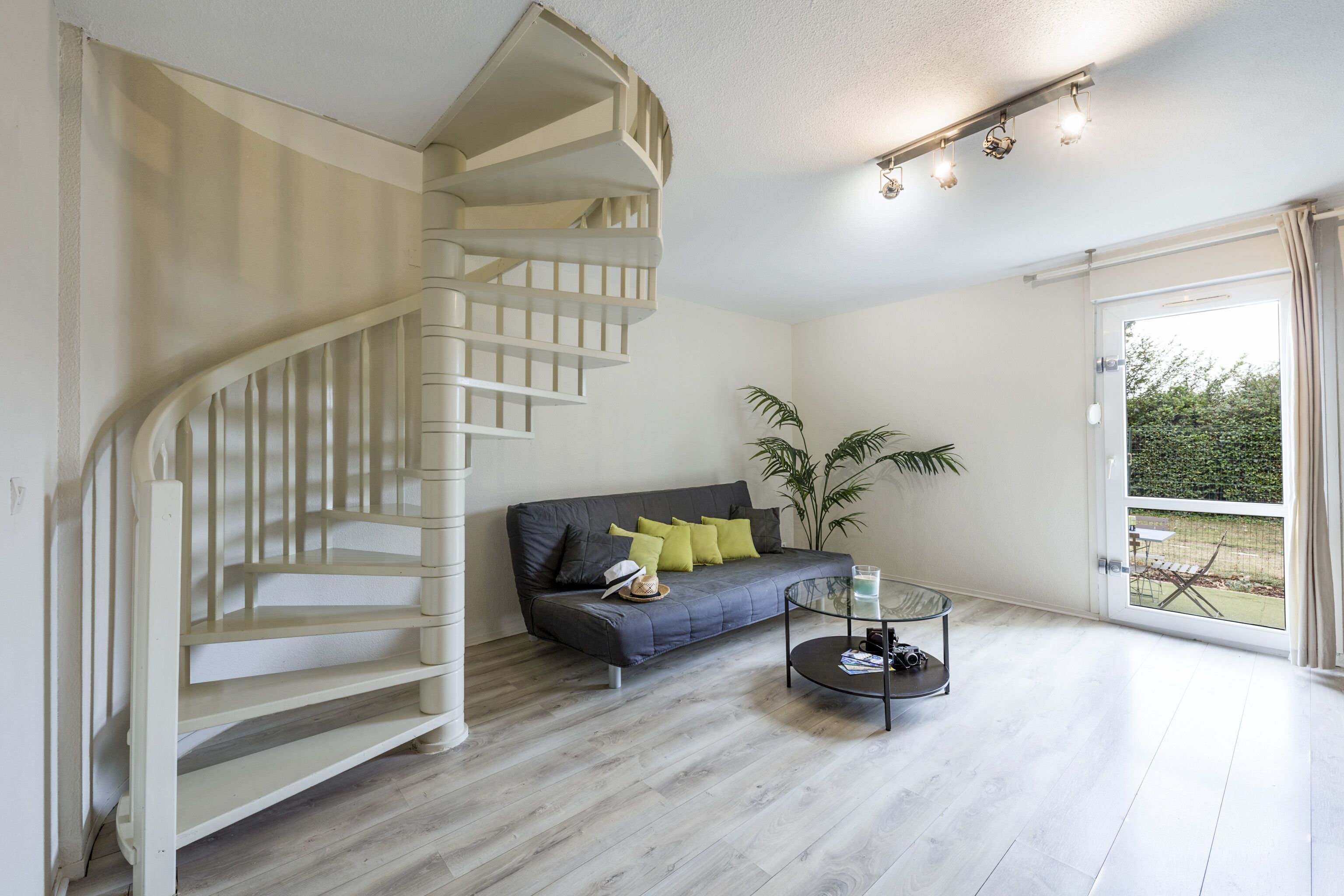 Una sala moderna con una escalera en espiral, un sofá gris con cojines, una mesa de centro de vidrio y vistas a la vegetación a través de un ventanal grande.