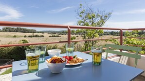 Una mesa con bebidas, aperitivos y un cuenco de comida en un balcón con vistas a un campo y árboles.