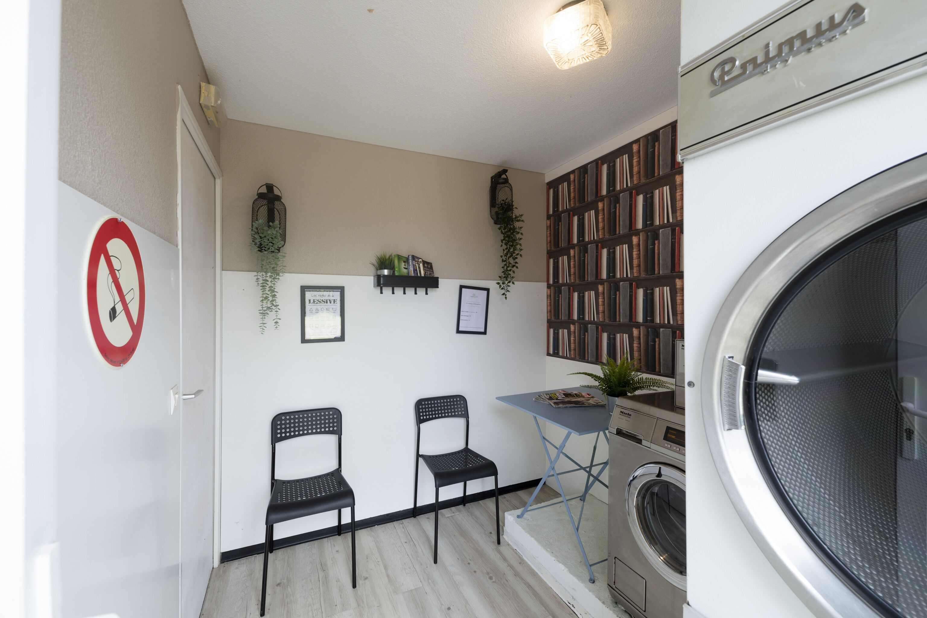 A compact laundry room with a washing machine, two chairs, a small table, and a bookshelf filled with books.