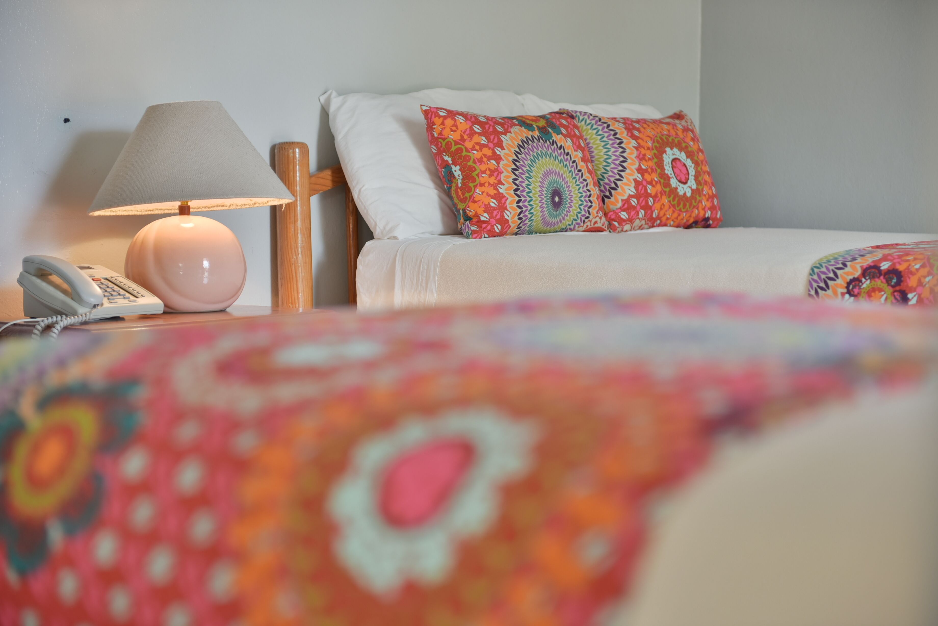 A neatly made bed with a colorful pillow, a bedside lamp, and a telephone on a wooden nightstand.