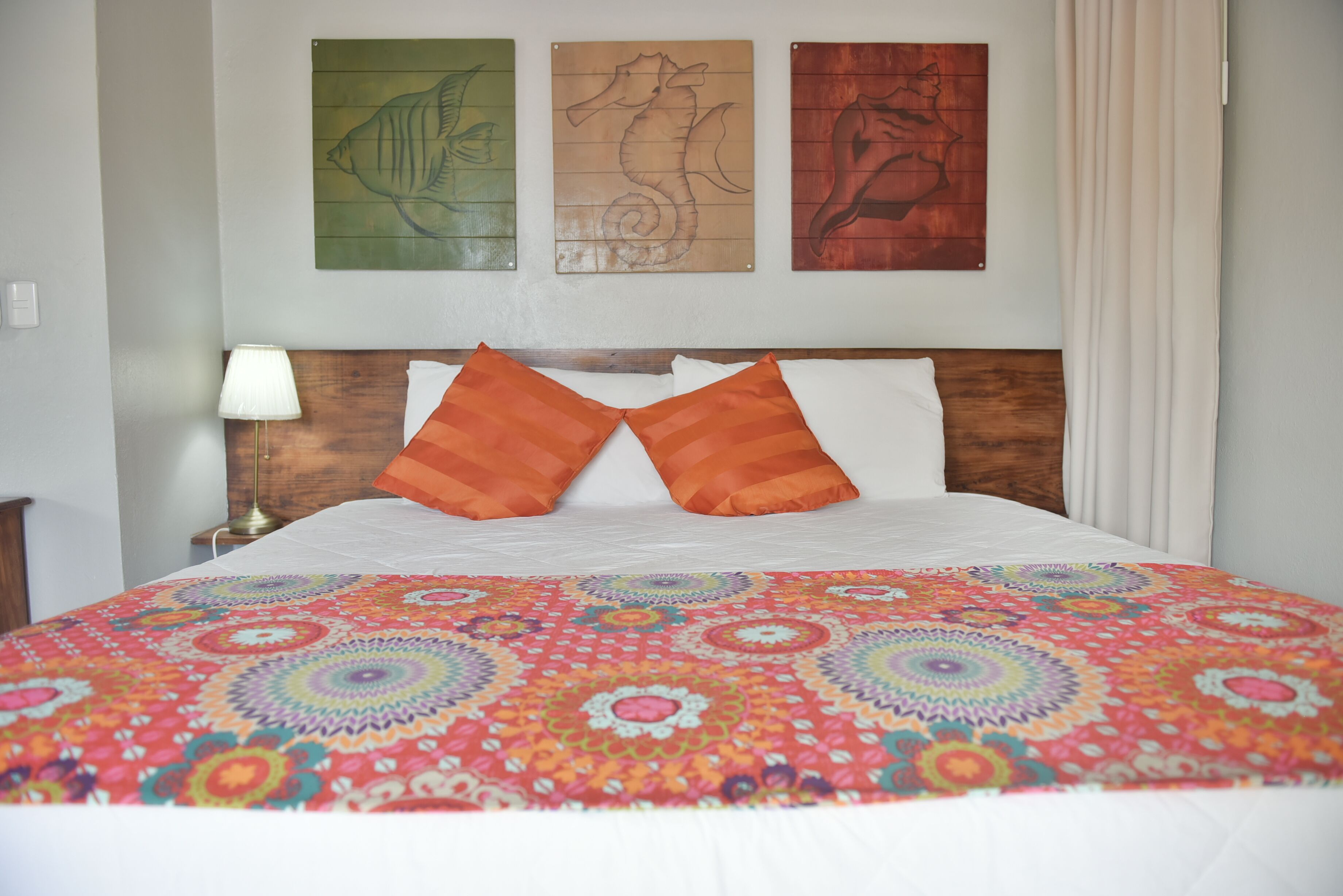 Habitación de hotel con cabecera de madera, colcha colorida y tres obras de arte en la pared que representan peces y un caballito de mar.